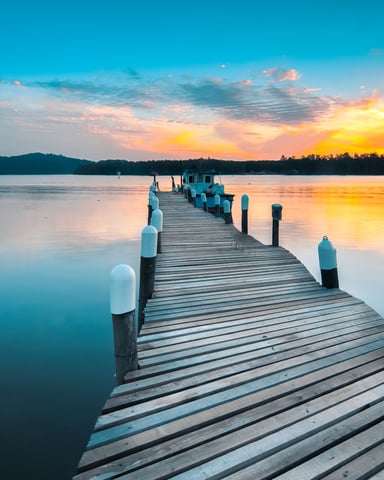 Waterfront marina with boats docked at wooden piers, blue water, and mountains in distance at sunset